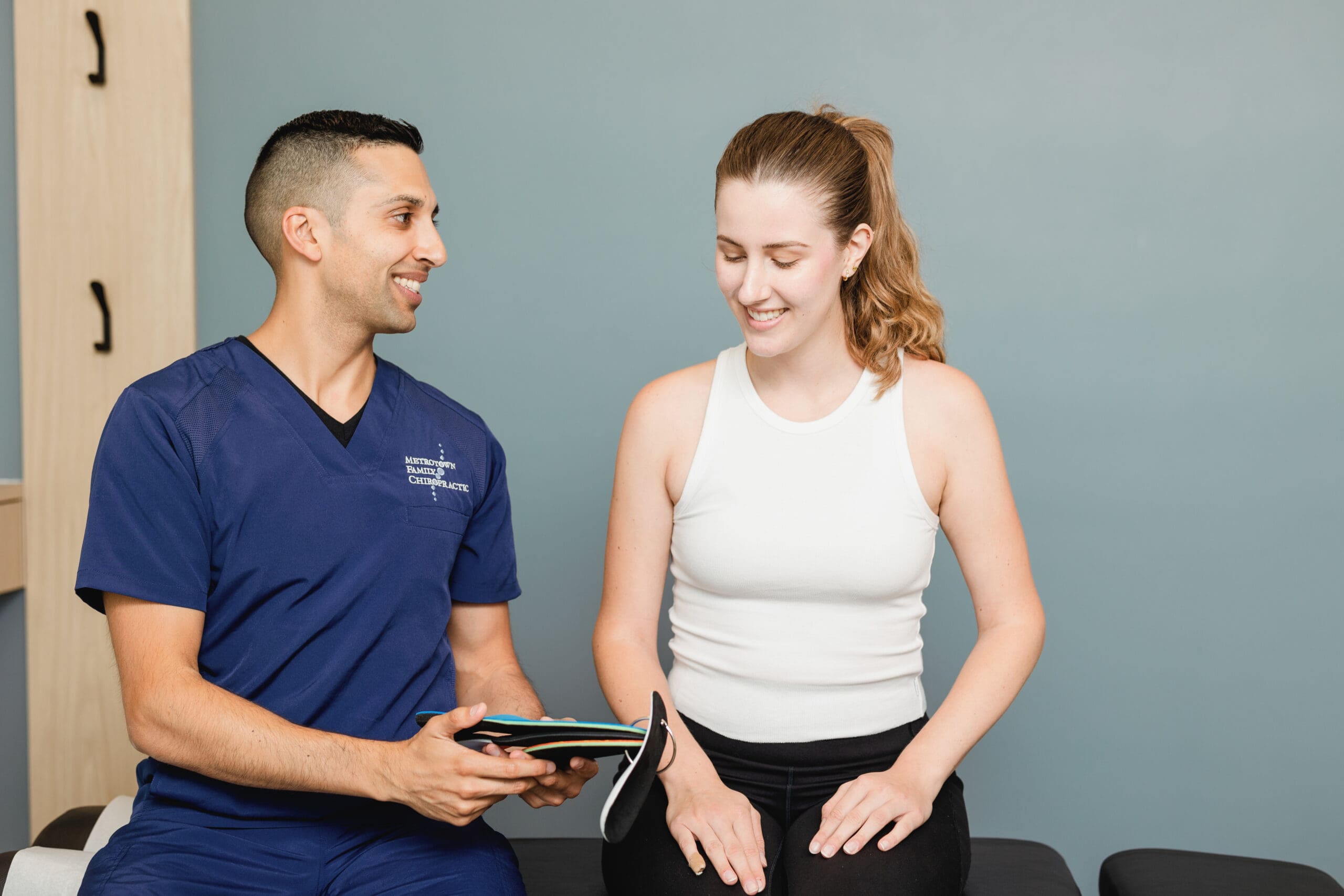 Chiropractor in blue scrubs holding a custom orthotic insole while smiling and discussing treatment options with a seated female patient. The patient, wearing a white tank top and black pants, is also smiling during the consultation. They are in a clinical setting with a neutral background.